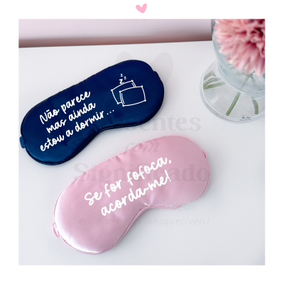 Máscaras de dormir azul escura e rosa claro com texto branco sobre uma superfície branca ao lado de um vaso com flores cor-de-rosa