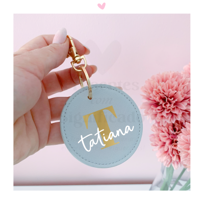 Chaveiro redondo cinza com letra T dourada e nome tatiana, segurado por mão com unhas cor de rosa, flores cor de rosa ao lado