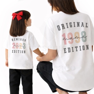 Camisolas brancas com estampados coloridos e texto, criança com laço vermelho no cabelo e mulher agachada ao lado.