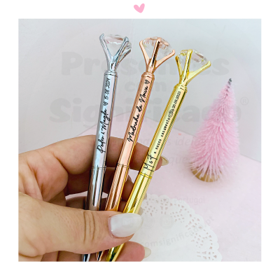 Três canetas metálicas com strass no topo seguradas por uma mão com unha branca, fundo com pinheiro rosa