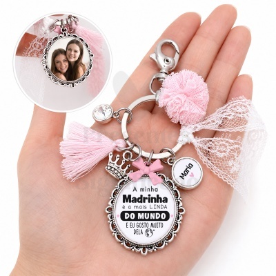 Chaveiro com pompom rosa, laço branco e medalhas com texto para madrinha, segurado por mão