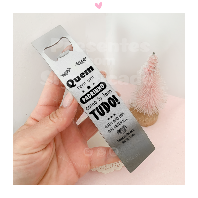 Abridor de garrafas metálico prateado com texto em português e desenho decorativo, sendo segurado por uma mão junto a uma pequena árvore decorativa rosa