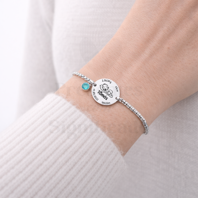 Pulseira prateada com pendente redondo gravado com dados de nascimento e uma pedra azul