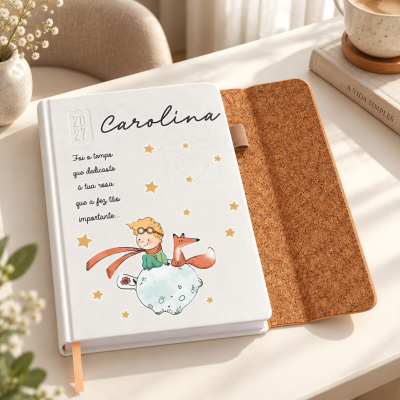 Agenda personalizada com capa branca e ilustração, aberta mostrando interior em cortiça, sobre mesa com vaso, café e livros.