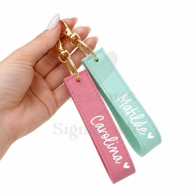 Dois porta-chaves retangulares rosa e verde água com nomes Carolina e Matilde em branco sendo segurados por uma mão.
