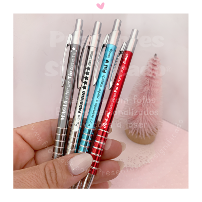 Quatro canetas metálicas com textos em português seguradas por uma mão, fundo rosa com mini árvore decorativa