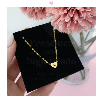 Colar dourado com pendente coração e letra M sobre cartão preto com flores cor-de-rosa ao fundo