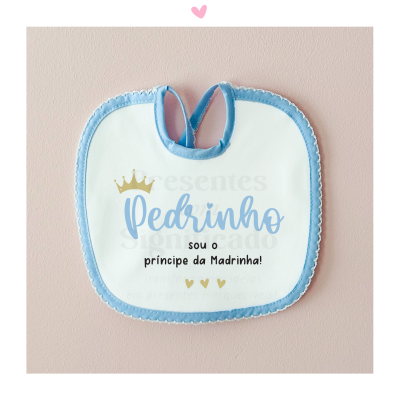 Babete de bebé branco com borda azul e texto 'Pedrinho sou o principe da Madrinha!' com coroa dourada em fundo rosa