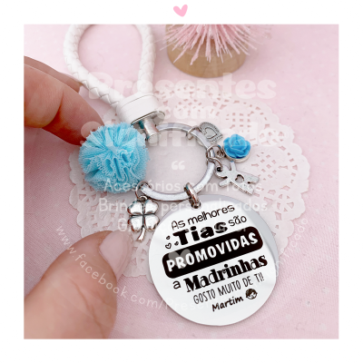Porta-chaves metálico prateado com texto em português, pompom azul e pequenas decorações, sobre fundo rosa e renda branca