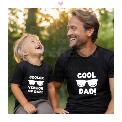 Homem e criança com camisolas pretas com textos 'COOL DAD!' e 'COOLER VERSION OF DAD!', sentados ao ar livre