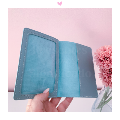 Carteira porta-cartões azul clara com costura branca numa mão, parede rosa e flores ao lado