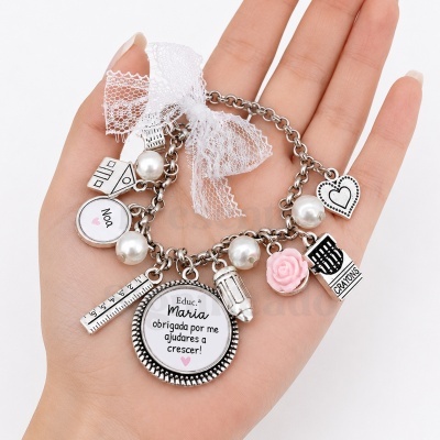 Pulseira com vários pendentes de metal e pérolas falsas com mensagem em português e laço de renda branca num fundo branco.