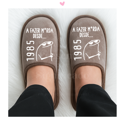 Chinelos castanhos com texto branco e ilustração, usados por pessoa de calças pretas sobre superfície branca felpuda