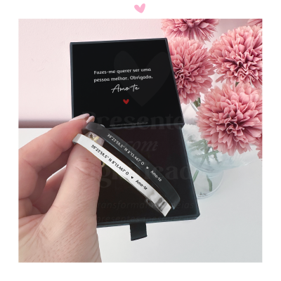 Pulseira prateada gravada com coordenadas e Amore-te em caixa preta com flores cor-de-rosa ao fundo
