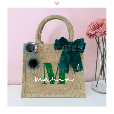 Saco de mão em ráfia bege com decoração verde e branca, laço verde escuro, óculos de sol azuis e flores cor de rosa.