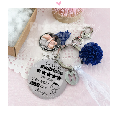 Chaveiro metálico com frases para madrinha, pompons azuis, fita branca, em superfície rosa e caixa ao lado.