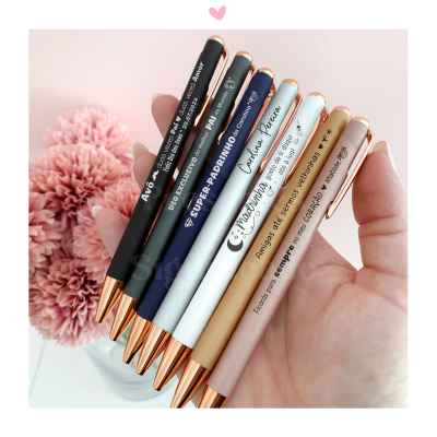 Conjunto de seis canetas metálicas coloridas com texto personalizado, seguradas por uma mão, flores cor-de-rosa ao fundo