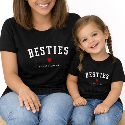 Mulher e menina a usar t-shirts pretas com texto BESTIES SINCE 2024 e coração vermelho, sentadas com jeans azuis