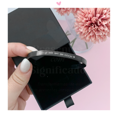 Pulseira preta metálica com gravação branca segurada sobre caixa preta, com flor rosa ao lado