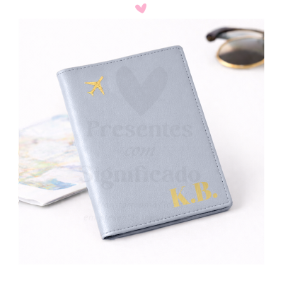 Estojo cinza claro com monograma dourado K.B. e avião dourado, mapa e óculos ao fundo.