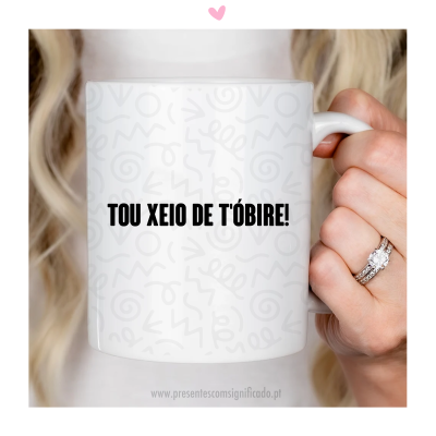 Caneca branca com texto 'TOU XEIO DE TÓBIRE!' e padrão cinza, segurada por mão com anéis