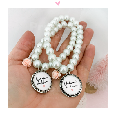 Pulseiras com pérolas brancas e pingentes escritos para madrinhas, seguradas numa mão.