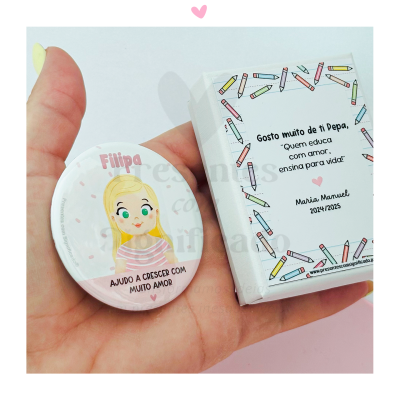 Botton redondo com ilustração de menina e texto em português, dentro de embalagem branca decorada com lápis coloridos