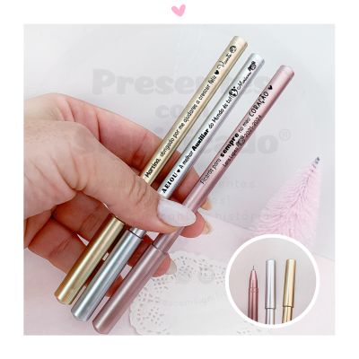 Três canetas metálicas dourada, prata e rosa ouro com texto preto sendo seguradas por mão com unhas brancas