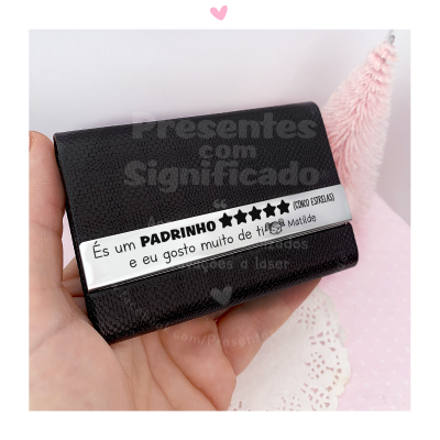 Carteira preta pequena com etiqueta branca personalizada segurada por mão sobre fundo claro com pano rendado e árvore rosa