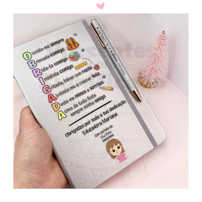 Caderno prateado com texto colorido e caneta prateada sobre fundo rosa com decoração de árvore pequena