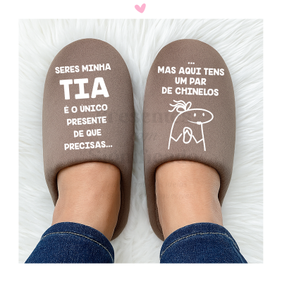 Par de chinelos castanhos com texto branco e desenho simples, usados por pessoa com jeans, sobre fundo branco felpudo.