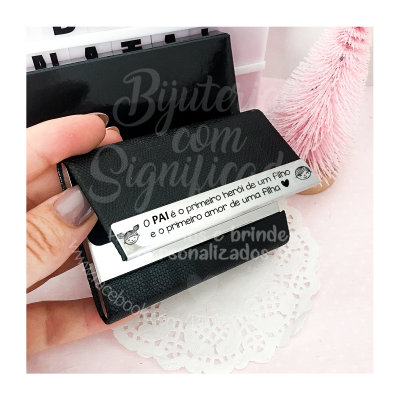 Etiqueta metálica negra com texto em português, mão segurando, fundo decorativo rosa e papel rendado branco