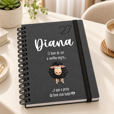 Caderno preto espiral com elástico, com design personalizado e figura de ovelha negra sobre mesa clara com café e planta.