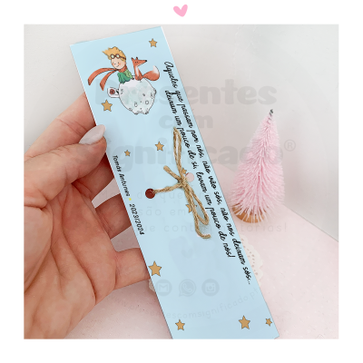 Marcador de página azul claro com ilustração do Pequeno Príncipe e texto em português, amarrado com fio de juta, sendo segurado por mão feminina, com mini árvore cor-de-rosa ao fundo.
