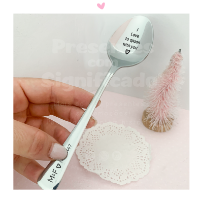 Colher metálica prateada com gravação de texto, fundo rosa e decoração