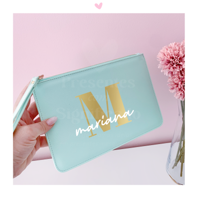 Carteira azul claro com letra M dourada e nome mariana em branco, segurada por mão, fundo rosa com flor