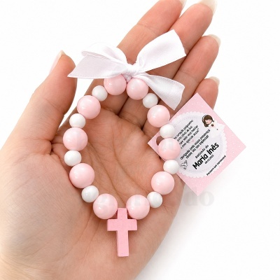 Pulseira com contas rosadas e brancas, crucifixo rosa e laço branco, com etiqueta cor-de-rosa para batizado