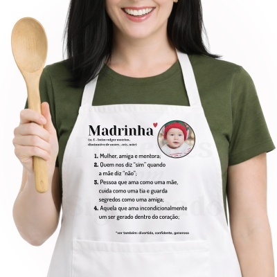 Avental branco com texto sobre madrinha e foto de bebé, usado por mulher com camisola verde, a segurar uma colher de madeira.