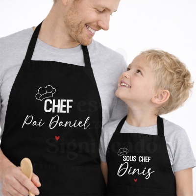 Aventais de cozinha pretos com texto CHEF Pai Daniel e SOUS CHEF Dinis usados por homem e criança.