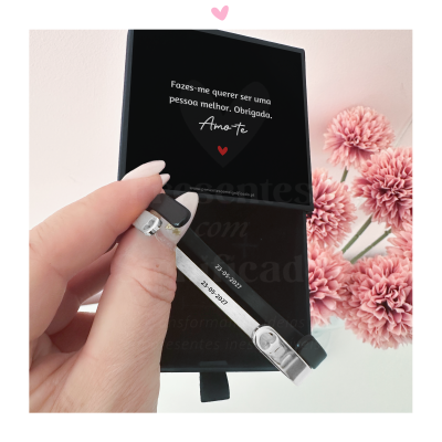Pulseira prateada com duas datas inscritas, segurada por mão com unhas brancas, quadro preto com texto e flores cor-de-rosa ao fundo