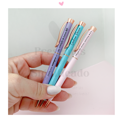 Três esferográficas coloridas com textos em português seguradas pela mão sobre fundo branco e rosa