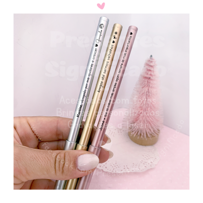 Três canetas metálicas coloridas com texto seguradas por uma mão em frente a uma árvore de Natal rosa