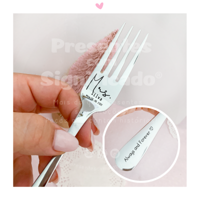 Garfo pequeno de metal prateado com texto gravado e fundo branco e rosa