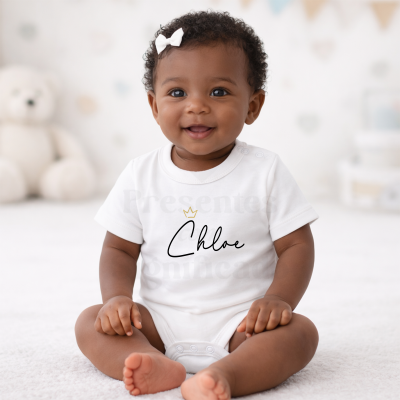 Bebé a sorrir com body branco escrito Chloe e coroa dourada sentado em tapete claro