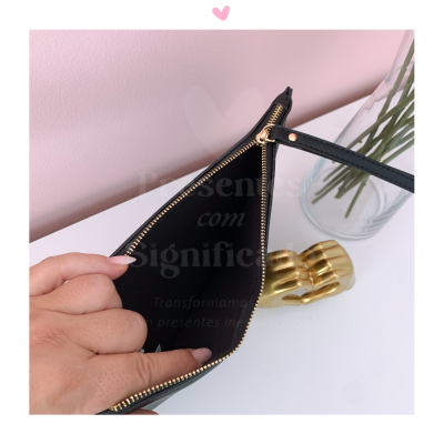 Carteira preta com fecho dourado aberta exibindo interior, objeto dourado e vaso de plantas num fundo rosa.