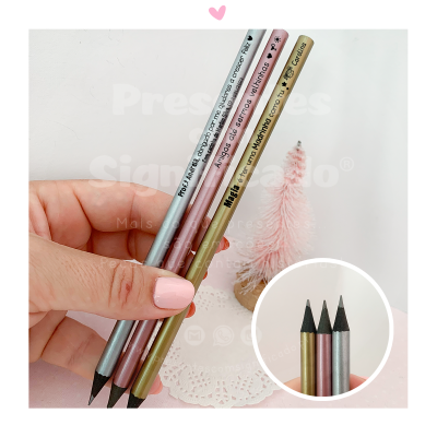 Lápis de cor metálicos rosa, cinzento e dourado com texto, em mão com unhas cor-de-rosa e pequena árvore cor-de-rosa ao fundo