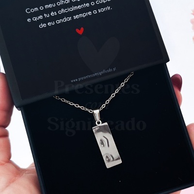 Colar com pendente retangular em metal prateado com imagem de olhos dentro de caixa preta com mensagem em português e coração vermelho.