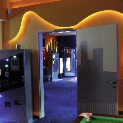 Porta branca com padrão cinza, parede amarela iluminada, máquina Coin Recycler cinza, mesa de bilhar verde