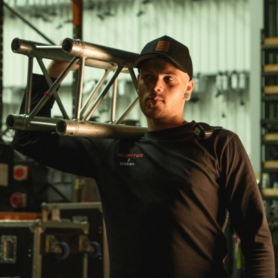 Homem com camisola preta e boné carregando estrutura metálica em ambiente industrial