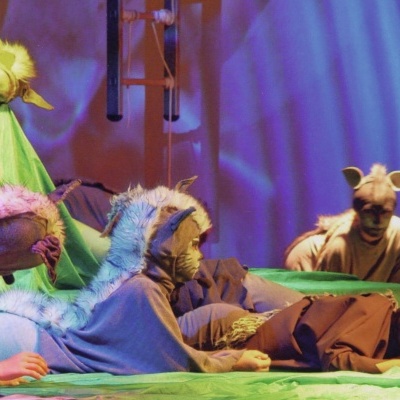 Atores em fantasia de animais numa cena teatral num palco com fundo azul e elementos verdes.